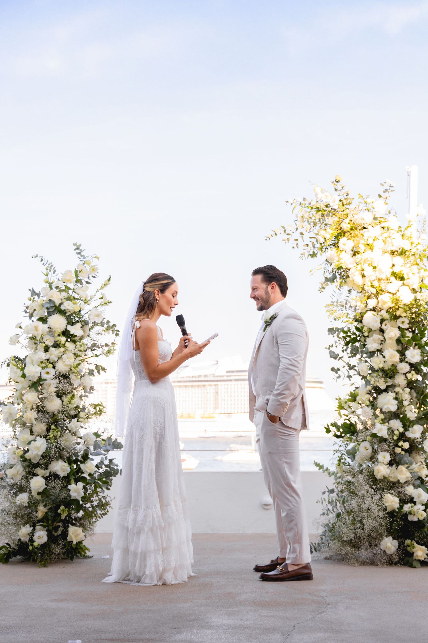 A wedding ceremony under white floral arches with a couple exchanging vows against a bright sky.