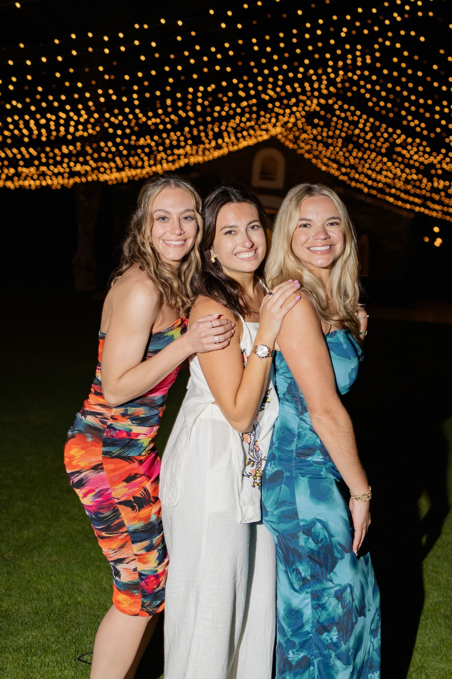 Three friends in colorful dresses pose together under twinkling string lights at an outdoor evening event.