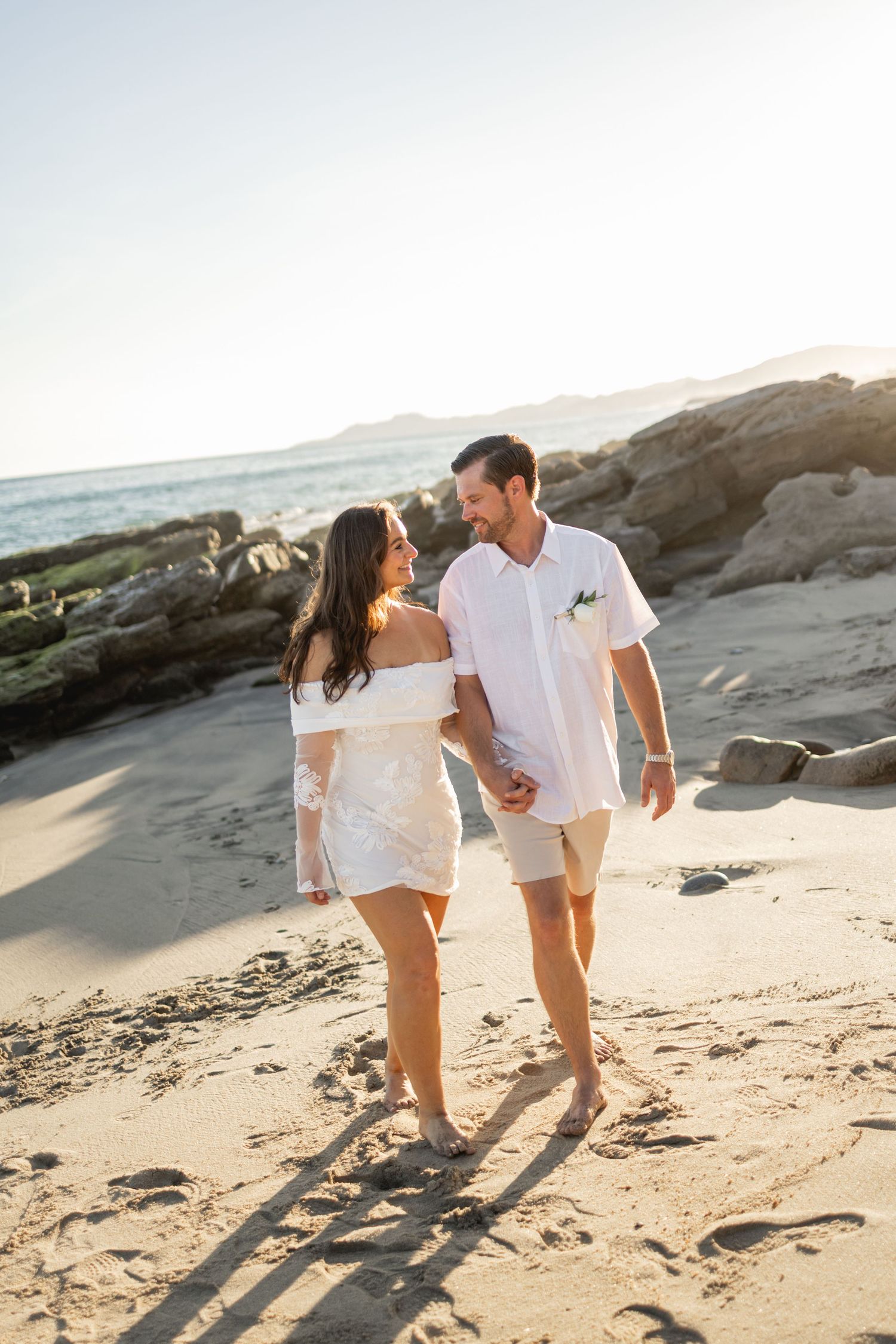 Couple walking along rocky beach shore in coordinated white outfits.