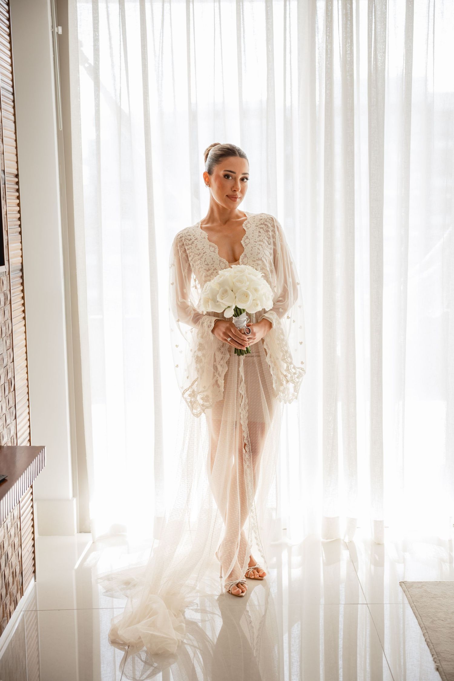 Elegant bridal portraits in a bright room with white curtains showing a flowing lace robe and bouquet.