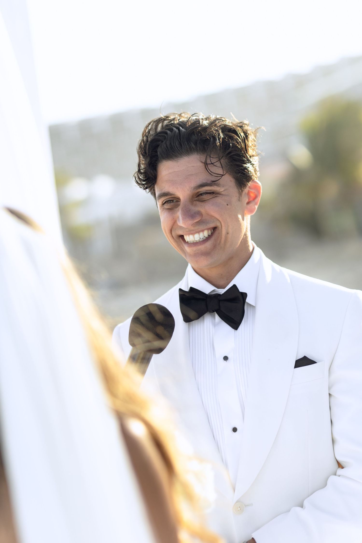 Close-up shots of groom in white tuxedo jacket and black bow tie smiling at beach wedding.