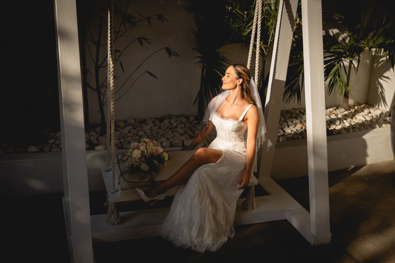 Bride in white dress sits elegantly on swing with dramatic lighting.
