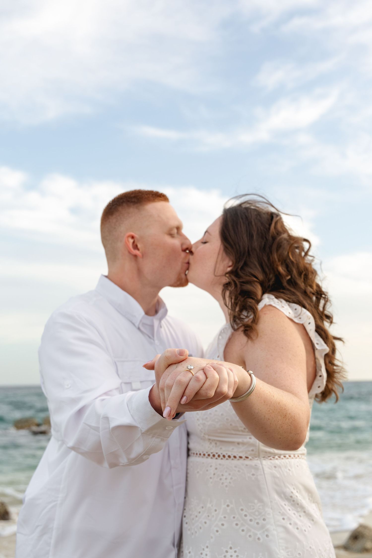 A romantic couple shares a tender kiss on a sunny beach with ocean waves in the background.