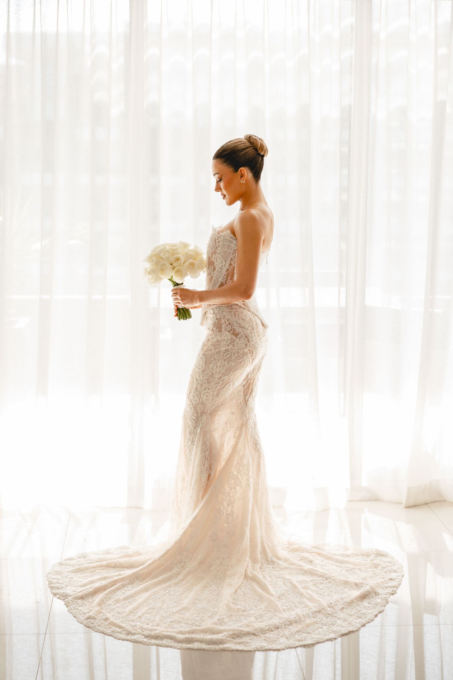 Elegant bride in a fitted lace wedding gown holding a white bouquet, standing before sheer curtains in soft natural light.