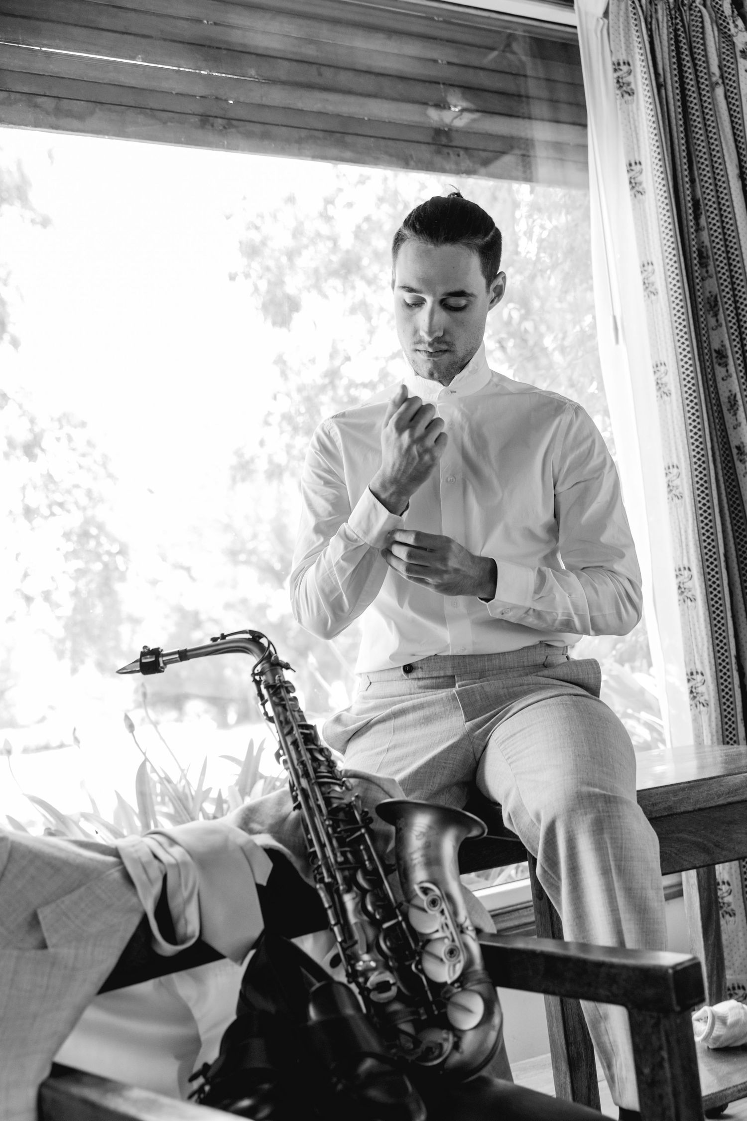 Musician in white attire taking a break during a saxophone session by a window.