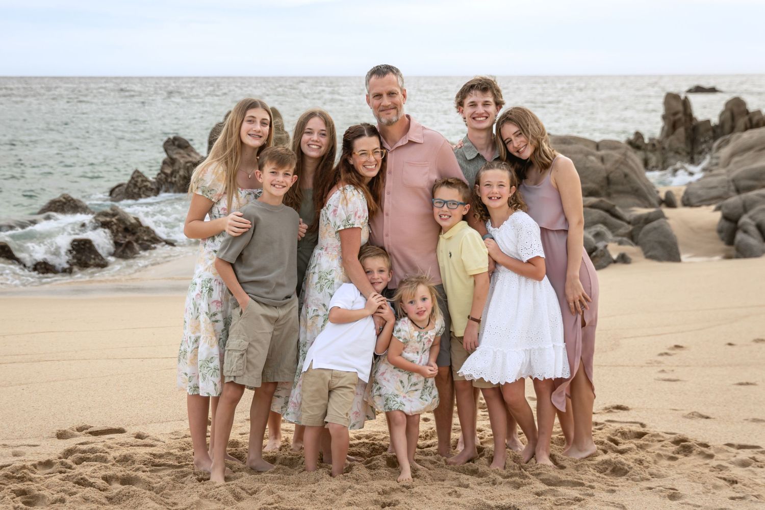 A group of people wearing casual beachwear pose together on a sandy beach with rocky coastline in the background.