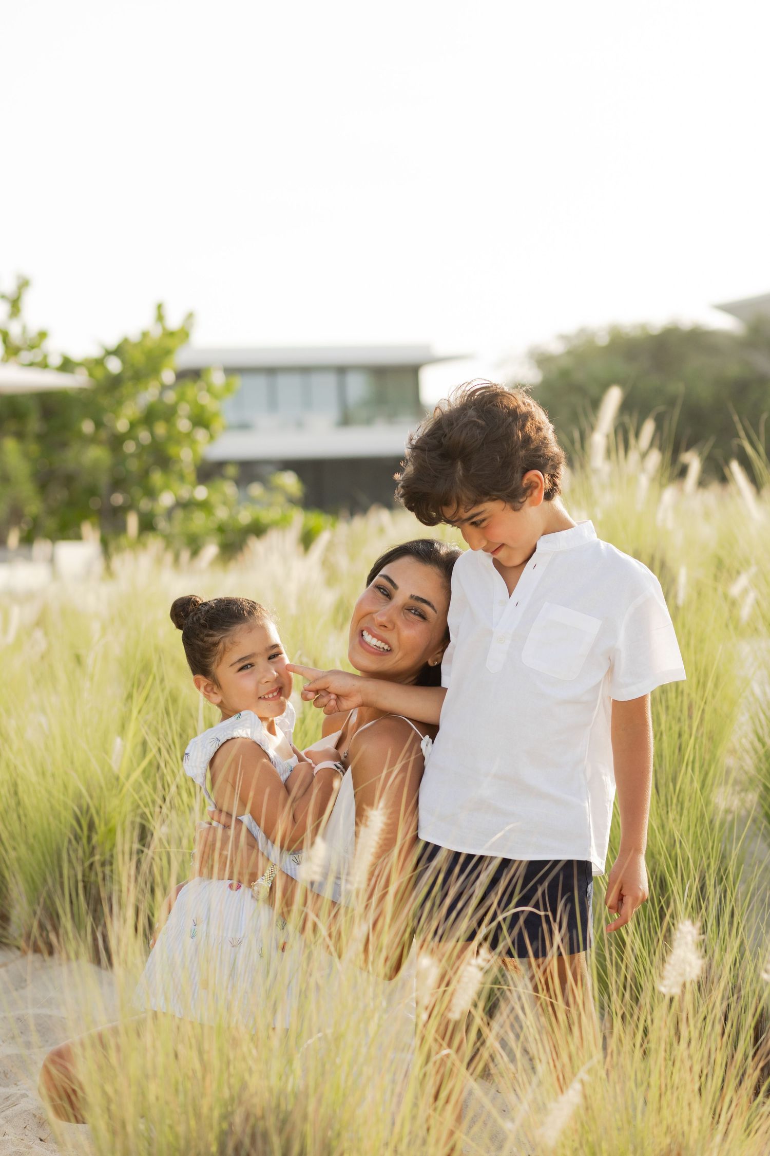 A smiling mother sits in tall grass with her two young children, a girl in white dress and a boy in white shirt, outdoors.