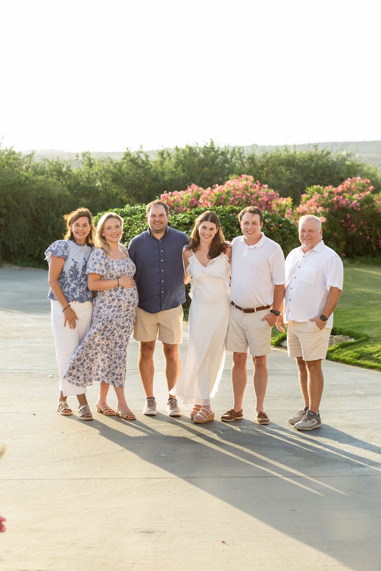 A group of people in casual summer attire stand together on a paved outdoor area casting long shadows at sunset.