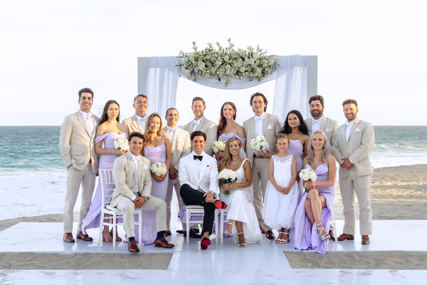 Wedding party in white and lavender attire gathered for formal beach ceremony photos.