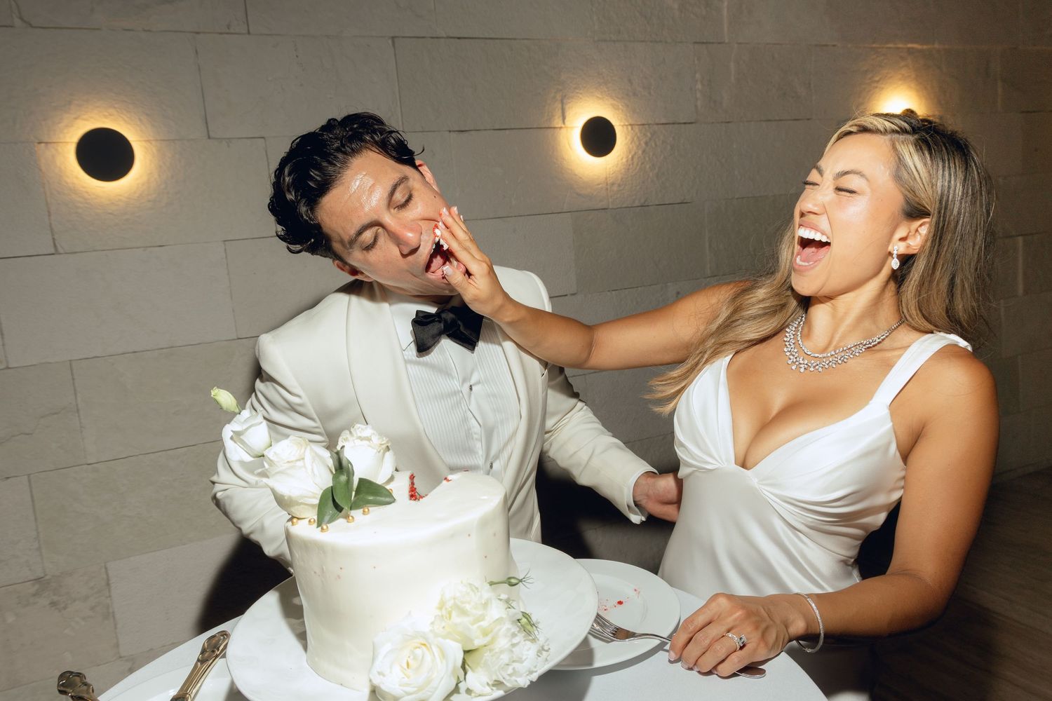 Newlyweds sharing a joyful cake-smashing moment against a concrete wall with sconce lighting.
