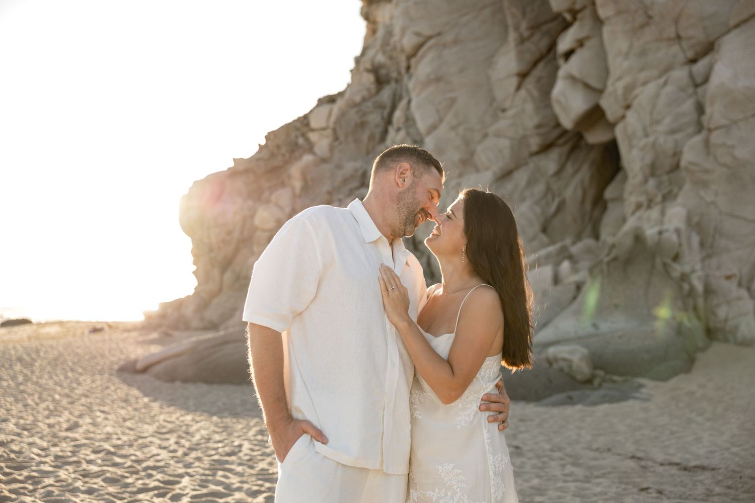 Couple in white outfits sharing a romantic moment on a sunlit beach with rocky cliffs in the background.