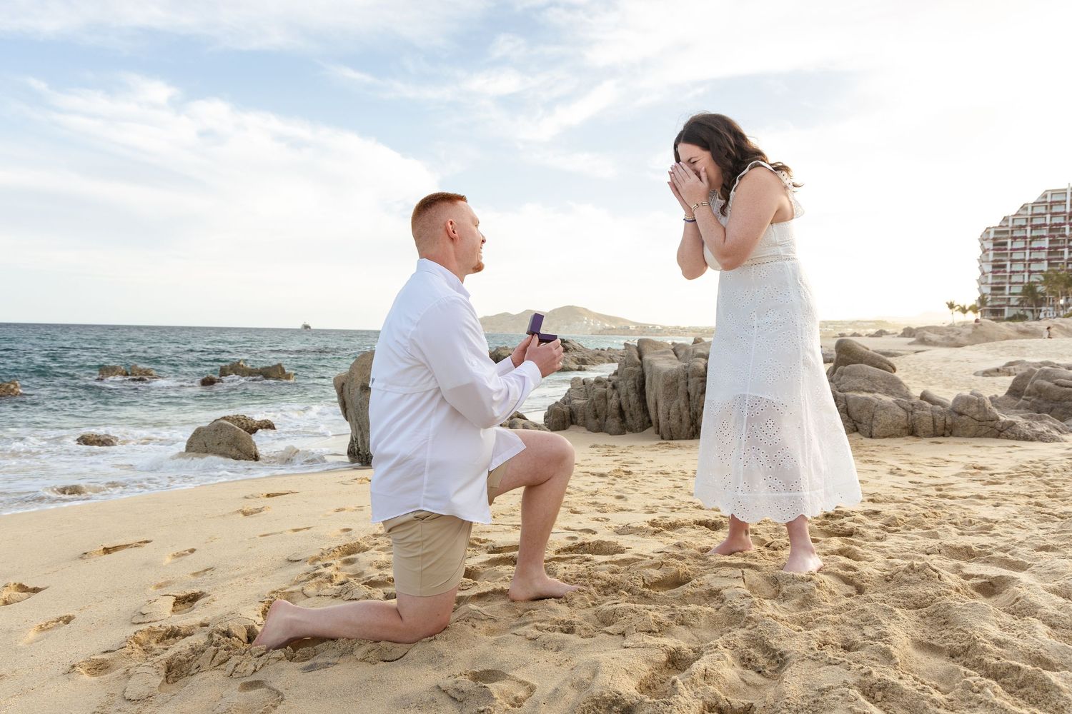 A romantic beach engagement photo session showing a couple in white outfits sitting on the sand at sunset.