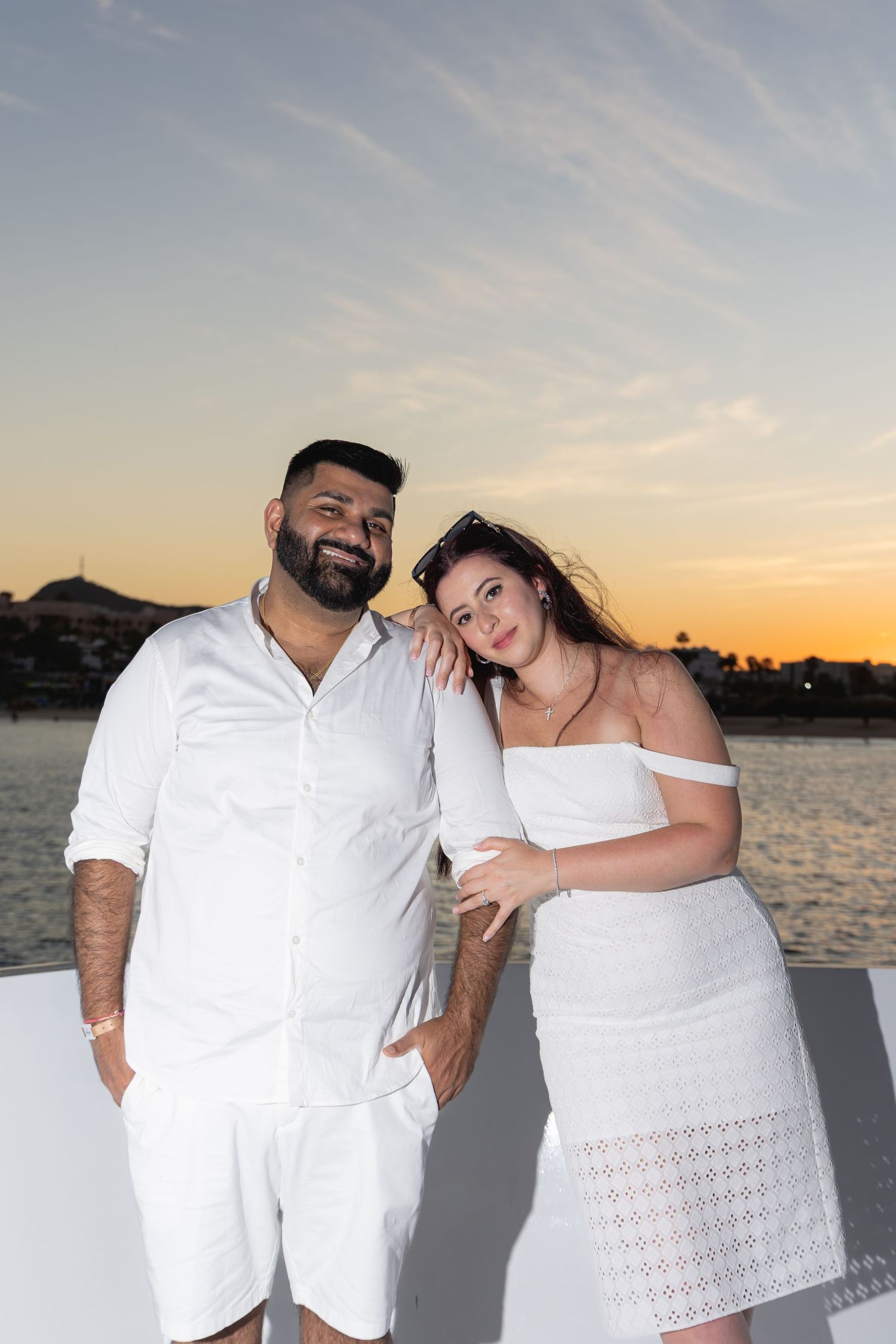 A couple in white outfits poses together for romantic sunset photos by the water.