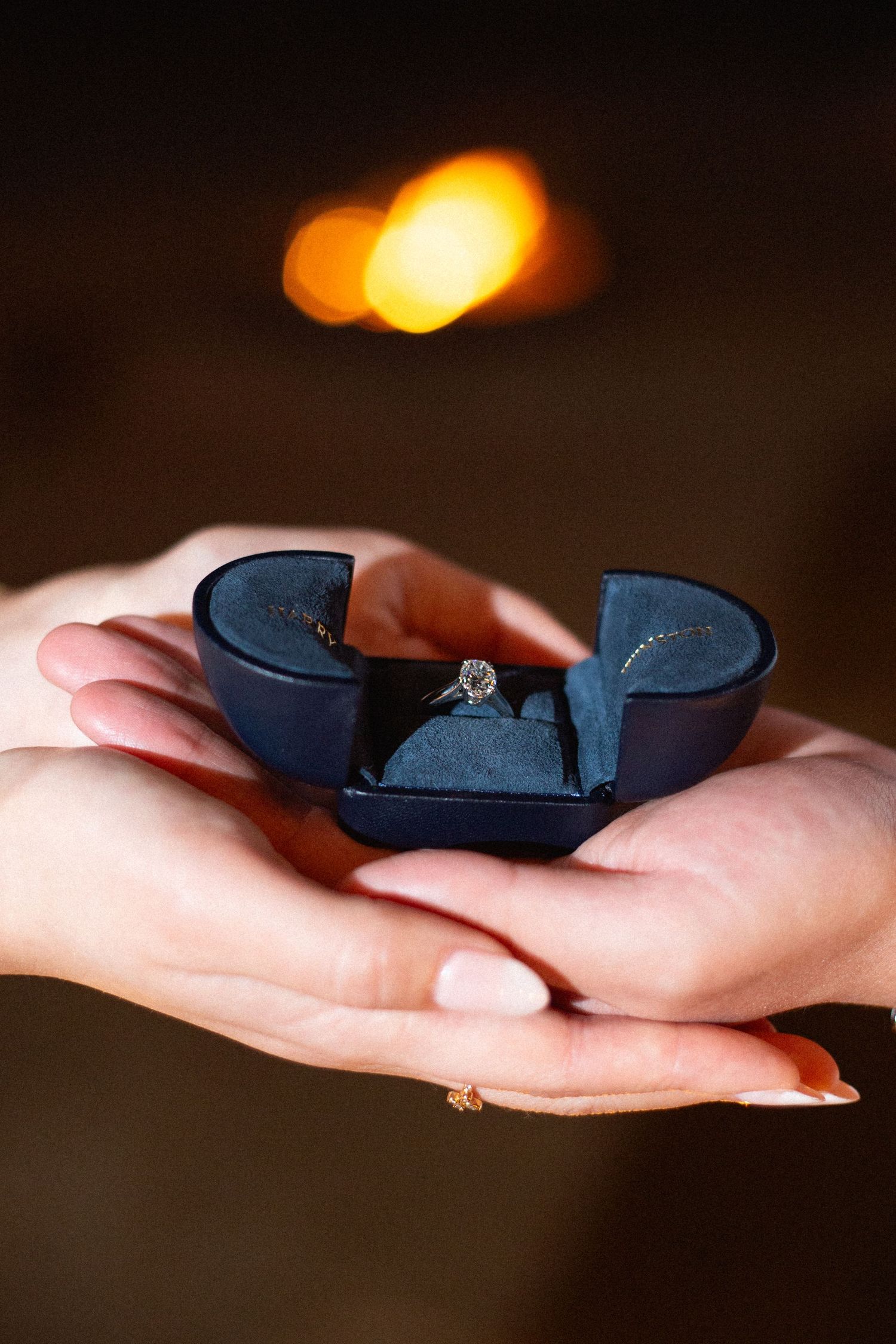 A hand holds a small navy blue ring box opening to reveal a sparkling diamond engagement ring in warm lighting.