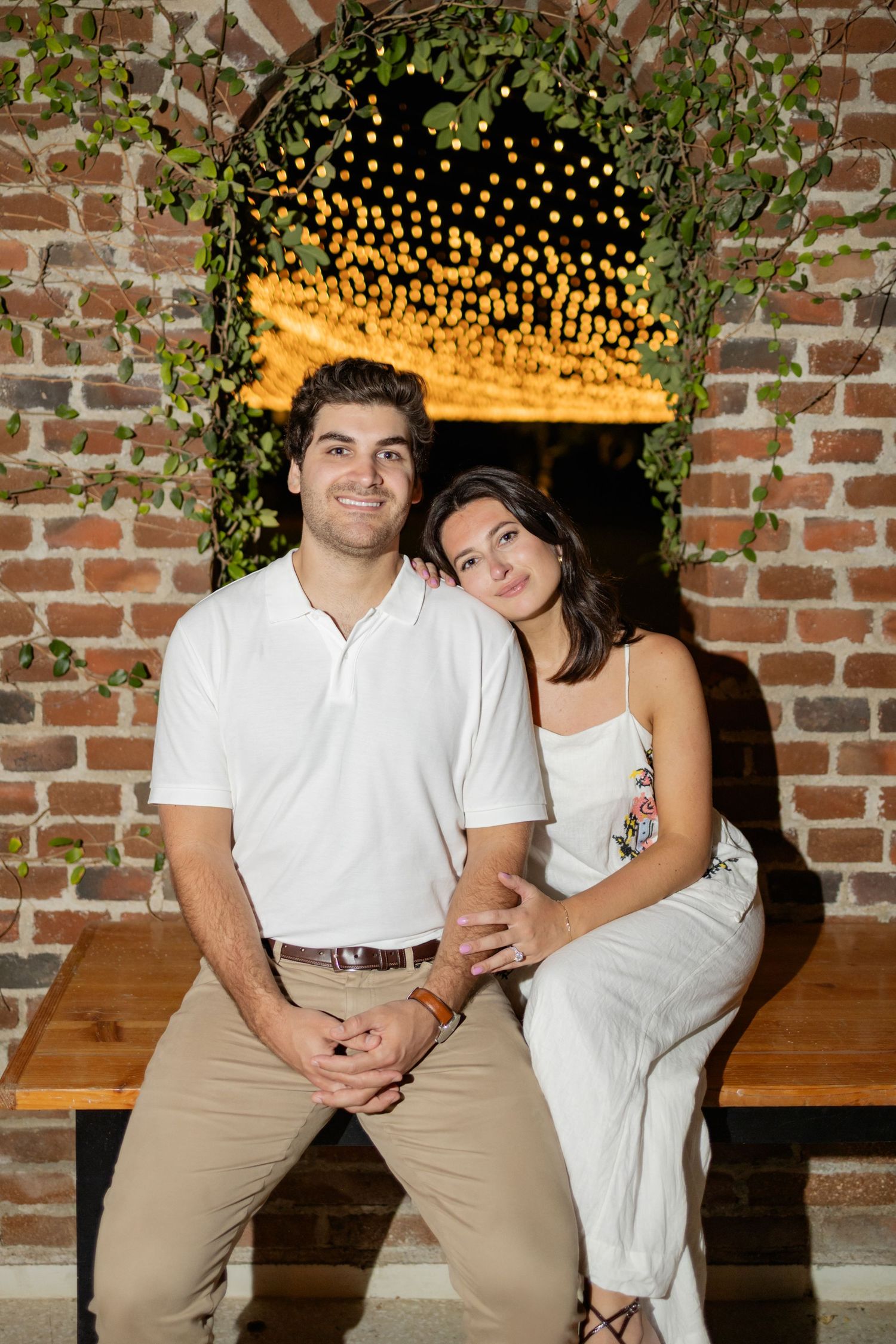 A couple in white shirts poses together against a brick wall with string lights and greenery decorations.