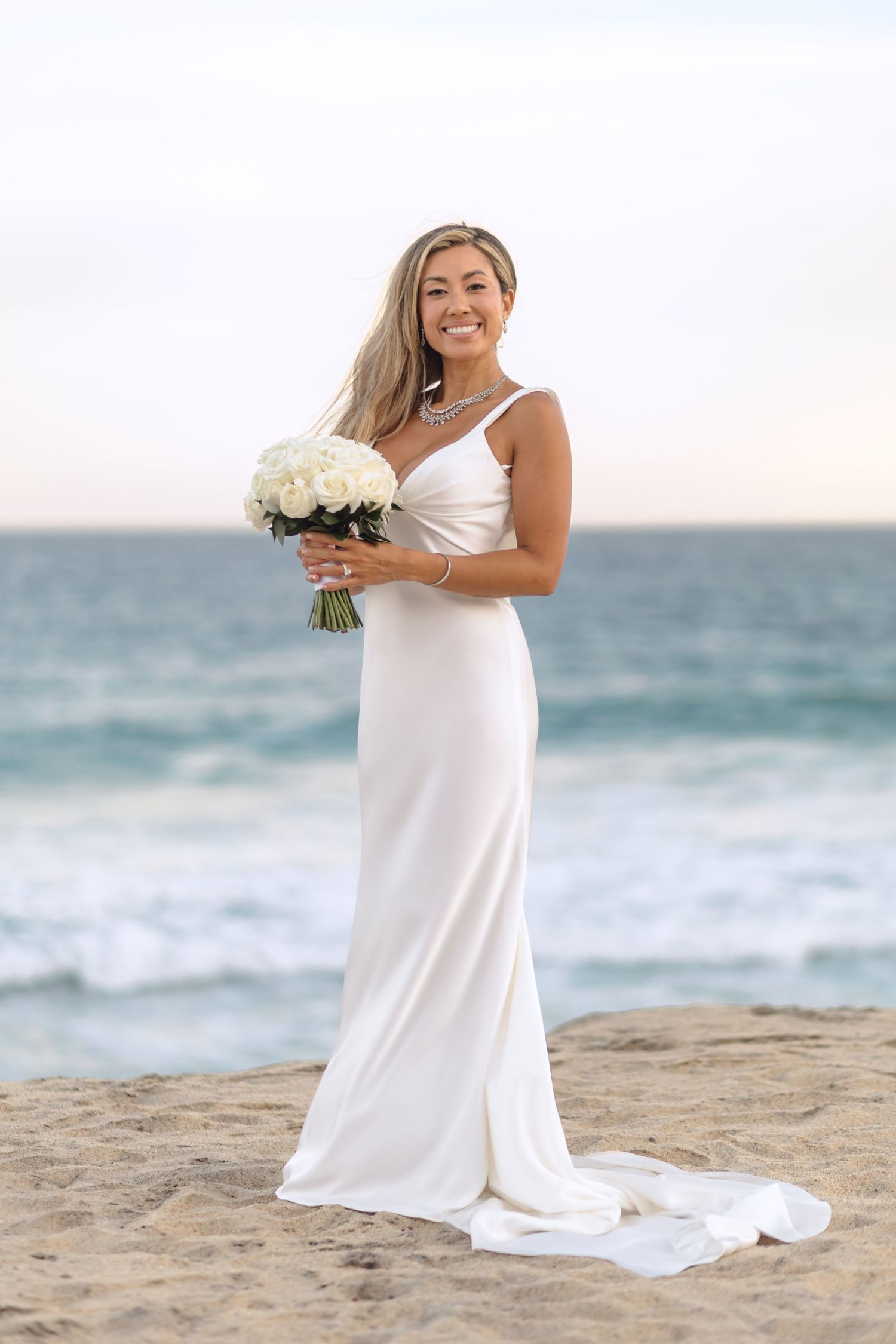 Bride in elegant white dress poses with bouquet on sandy beach with ocean backdrop.