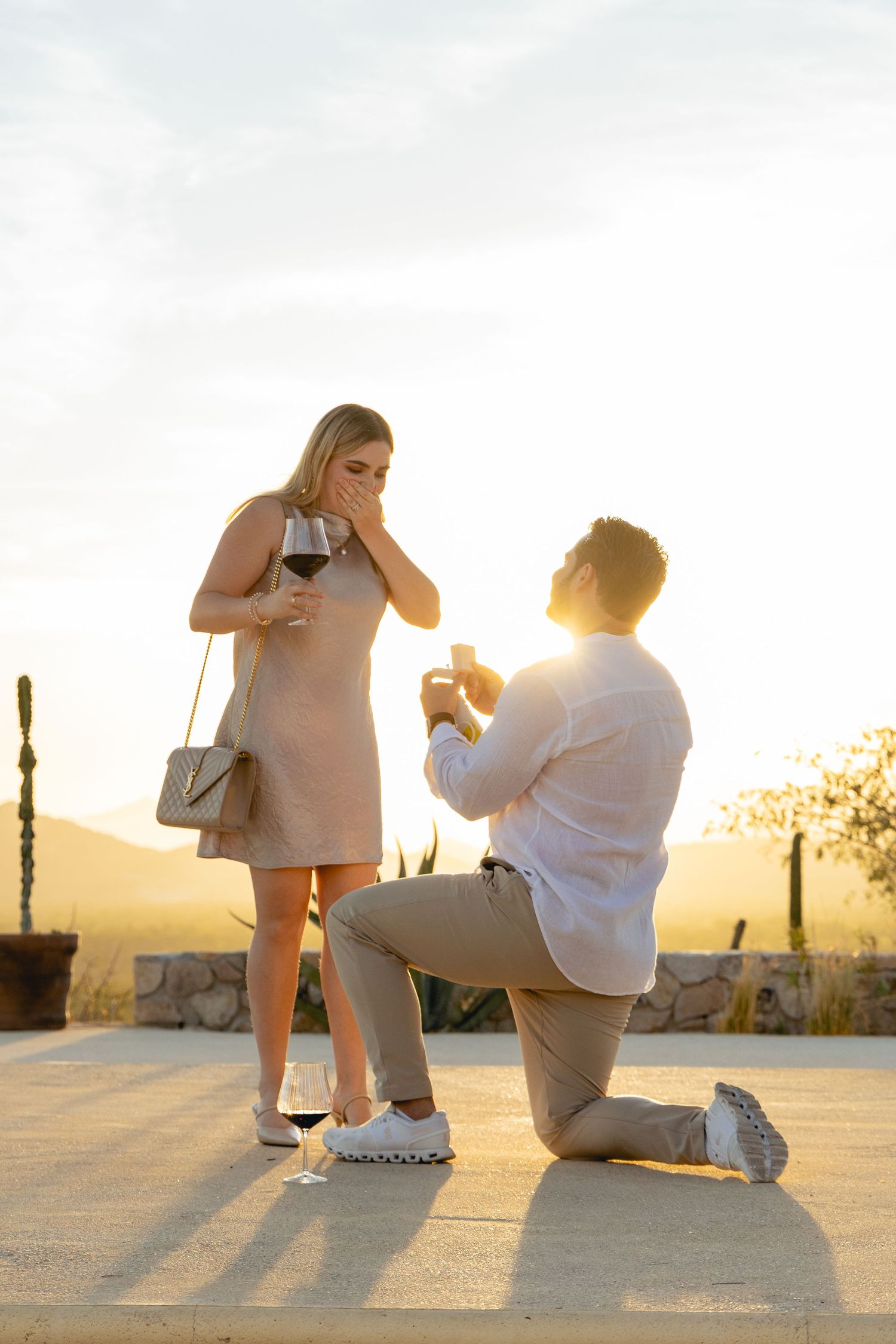 A couple in casual white outfits poses for a romantic sunset proposal photoshoot in a desert landscape.