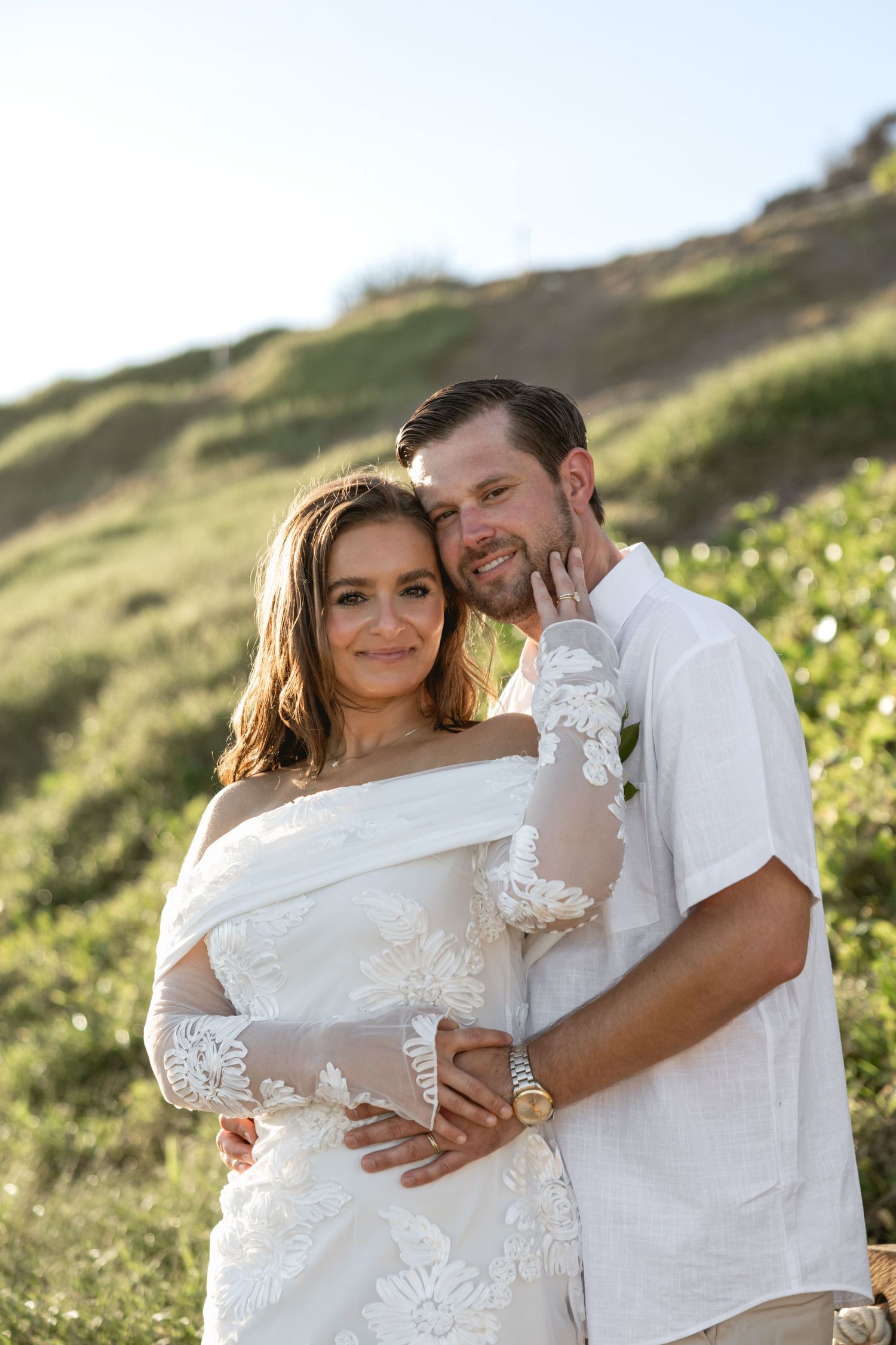 Couple embracing on grassy hillside during sunset photoshoot in white outfits.