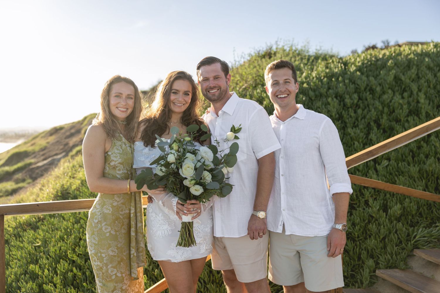 A group poses together in casual white and beige outfits against a natural coastal backdrop.