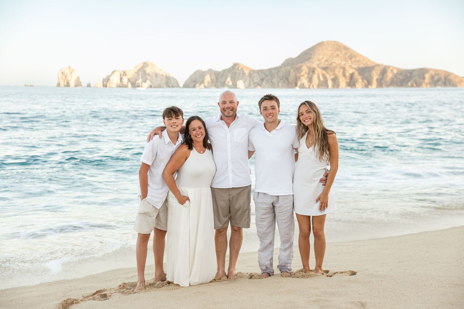 Family of five posing on a sandy beach in Cabo San Lucas with rocky cliffs and blue ocean in background.