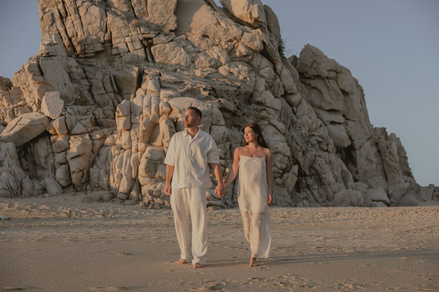 Couple in white attire holding hands on sandy beach with dramatic rocky cliffs at golden hour.