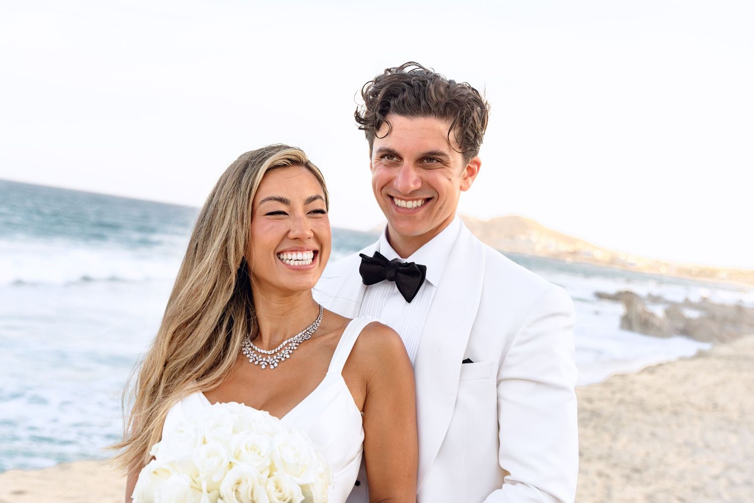 Romantic beach wedding portrait with couple in white attire against ocean backdrop.