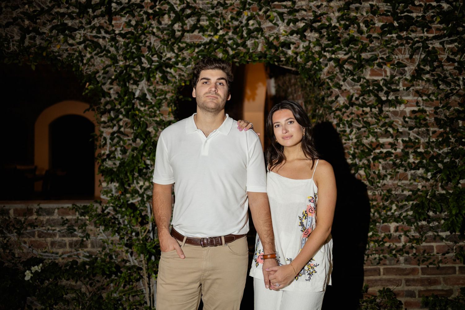 A couple poses together against an ivy-covered brick wall at dusk wearing white and khaki attire.