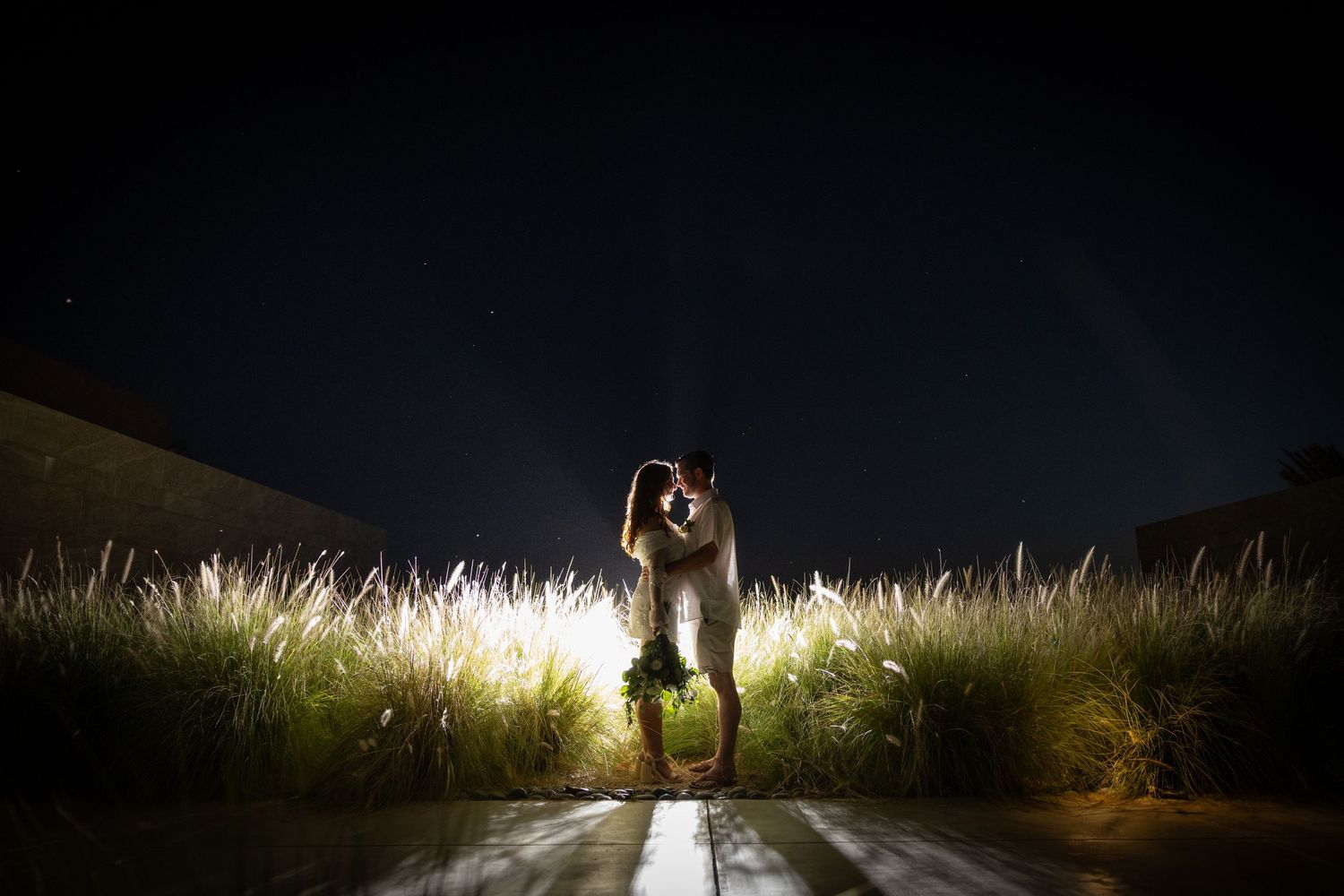 Couple in white outfits walking together at night with dramatic lighting and grass in the background.