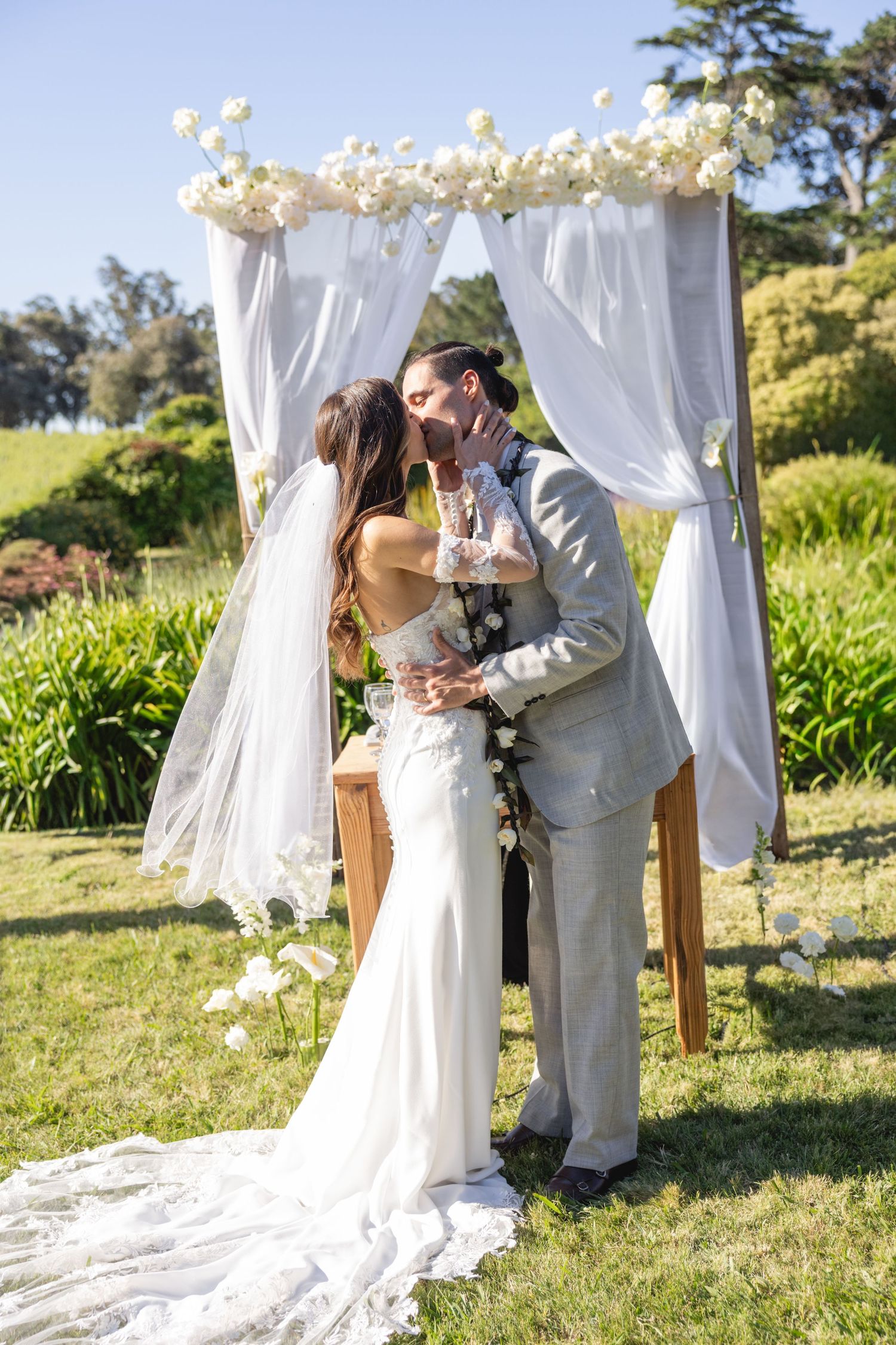 Wedding ceremony under white floral arch with flowing curtains in garden setting.