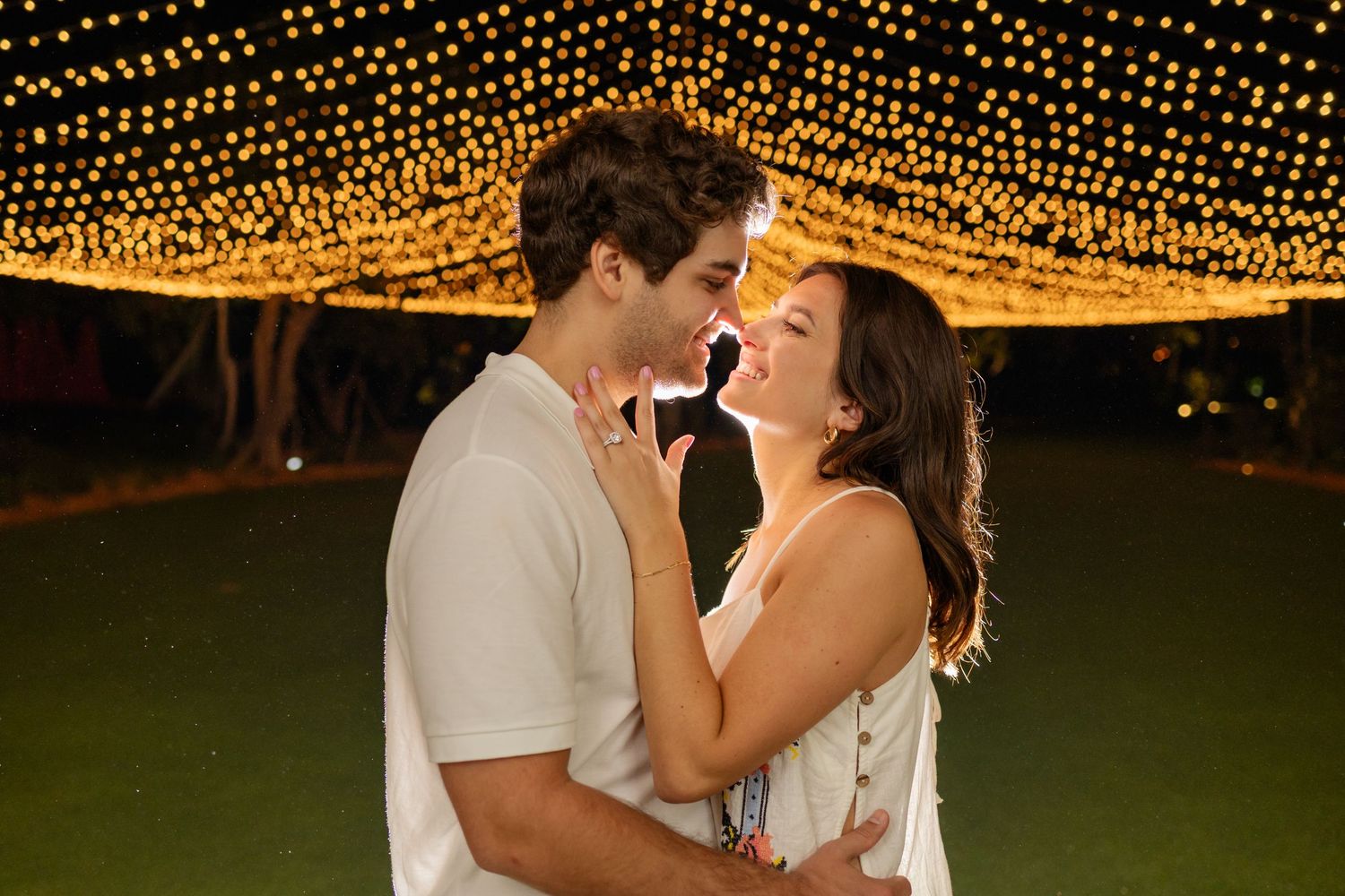 A romantic couple embraces under twinkling string lights at an outdoor evening celebration.