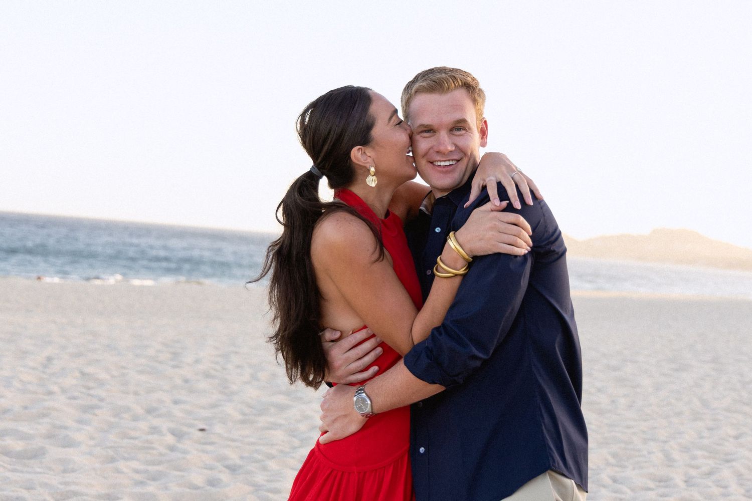 A couple embraces joyfully on a sandy beach at sunset wearing red and navy blue outfits.
