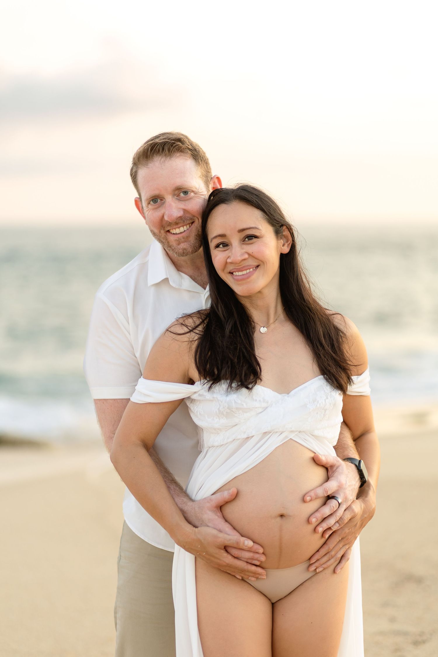 Beach maternity photoshoot featuring couple in white against sunset sky.