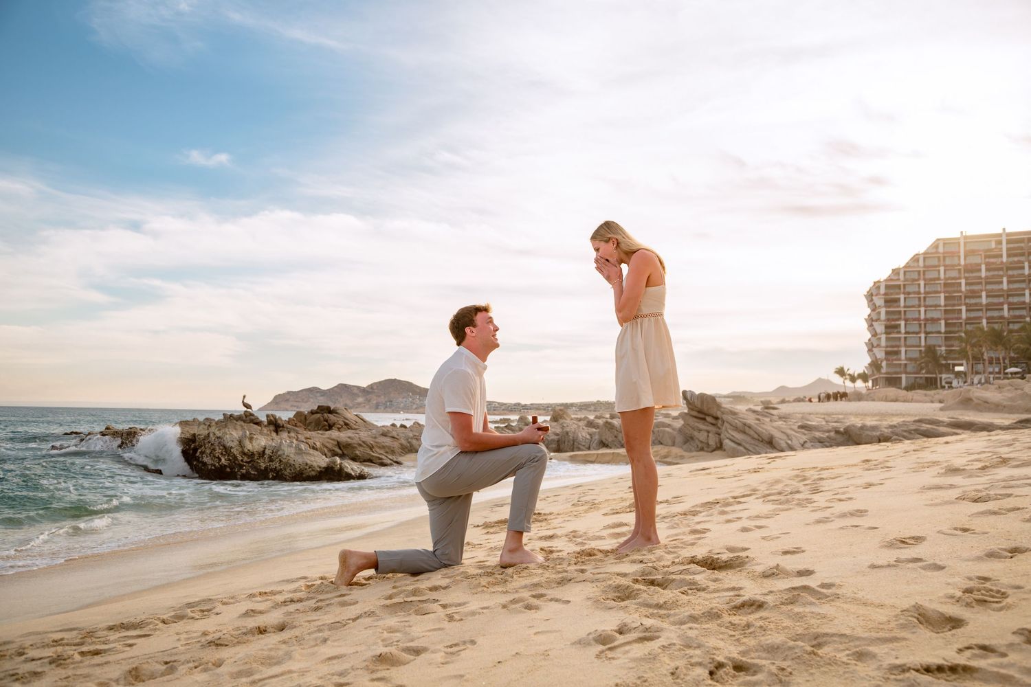 A romantic beach proposal scene unfolds at sunset with two figures silhouetted against the golden sky.