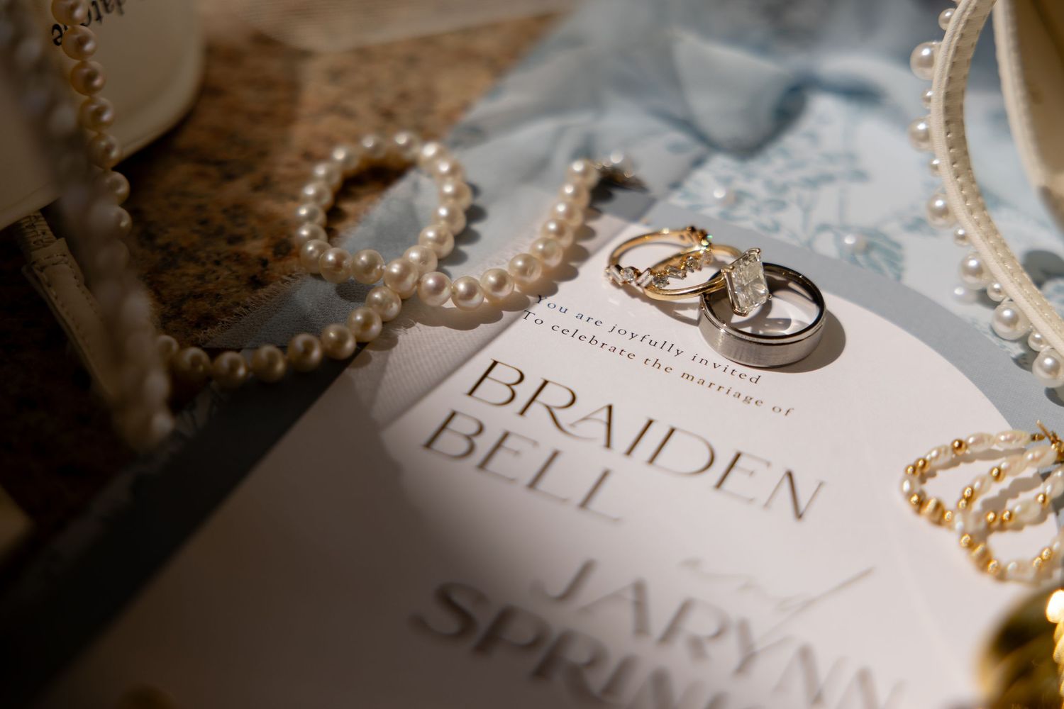 Wedding rings resting on invitation for Braiden Bell and Jarynn Spring, surrounded by pearl jewelry on soft blue fabric.