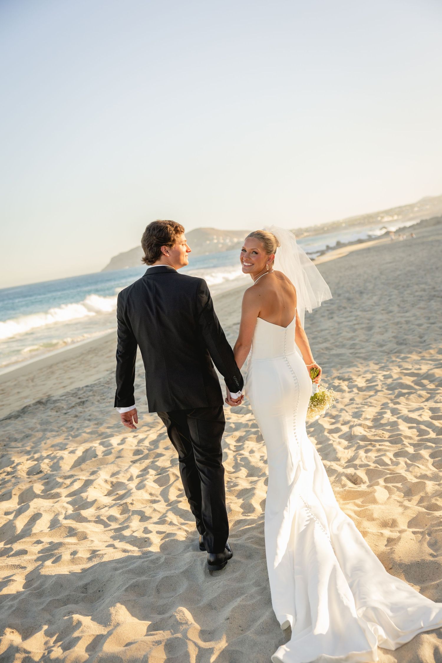 Newlywed couple walking hand in hand on a sandy beach, bride in white gown with veil, groom in black suit.