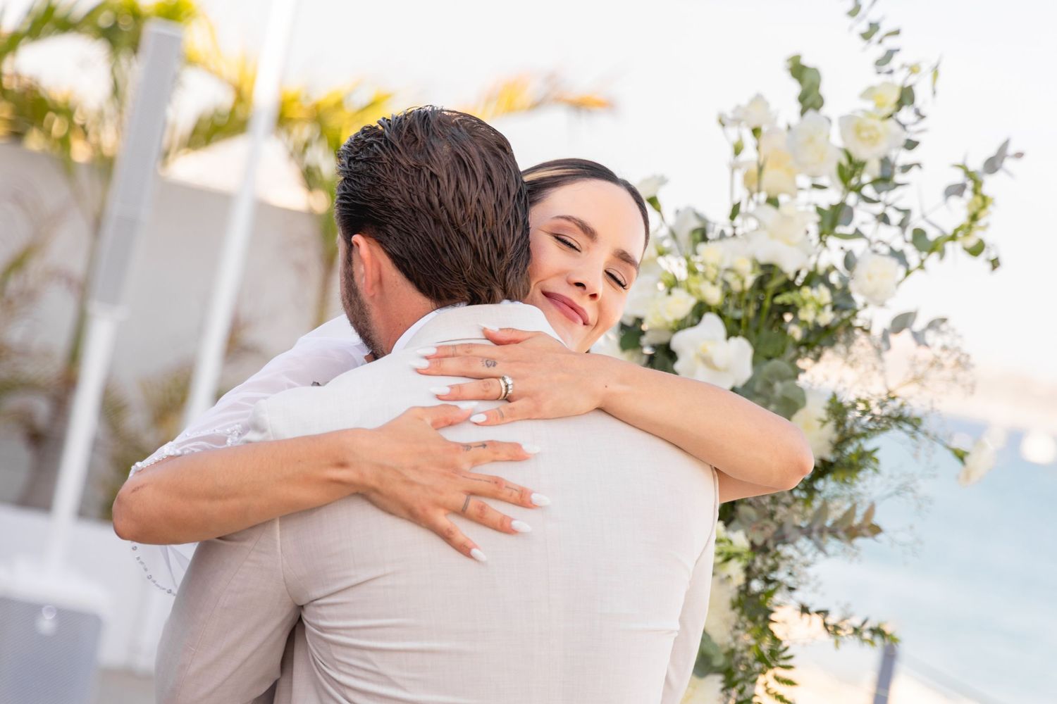 A couple embraces during an outdoor wedding ceremony with white floral arrangements in the background.