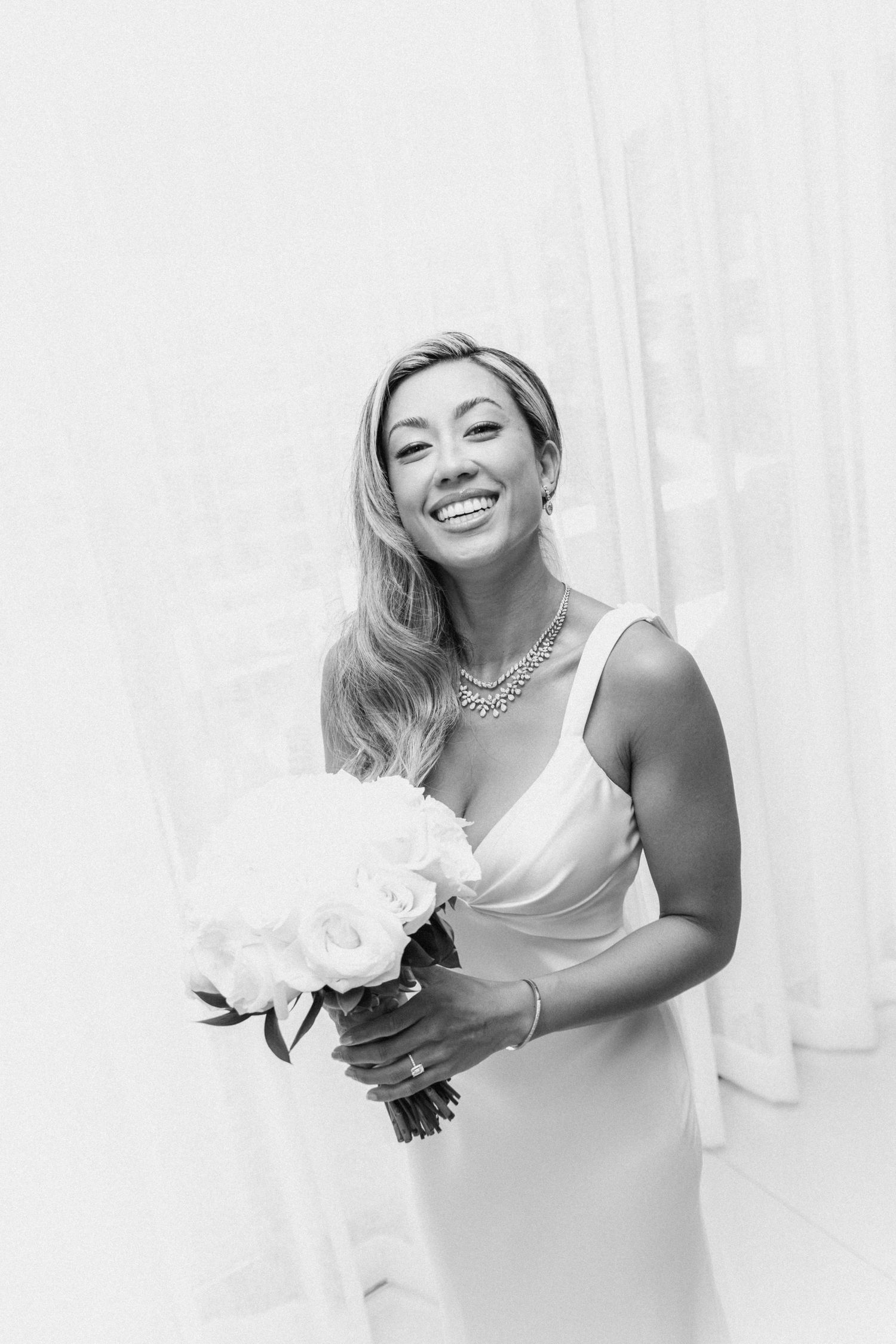 Bride in white dress holding bouquet of white flowers against bright background.
