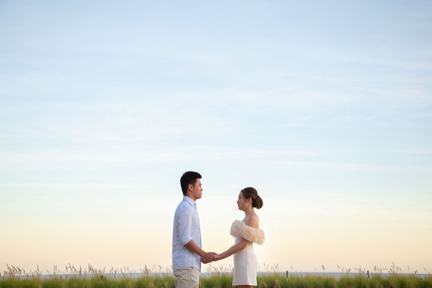 Two people in white clothing stand together on a grassy hill against a bright blue sky at sunset.