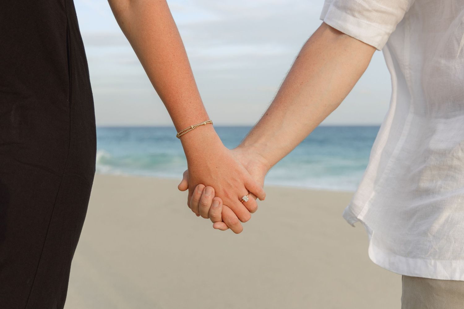 Two people hold hands while standing on a sandy beach with ocean waves in the background.