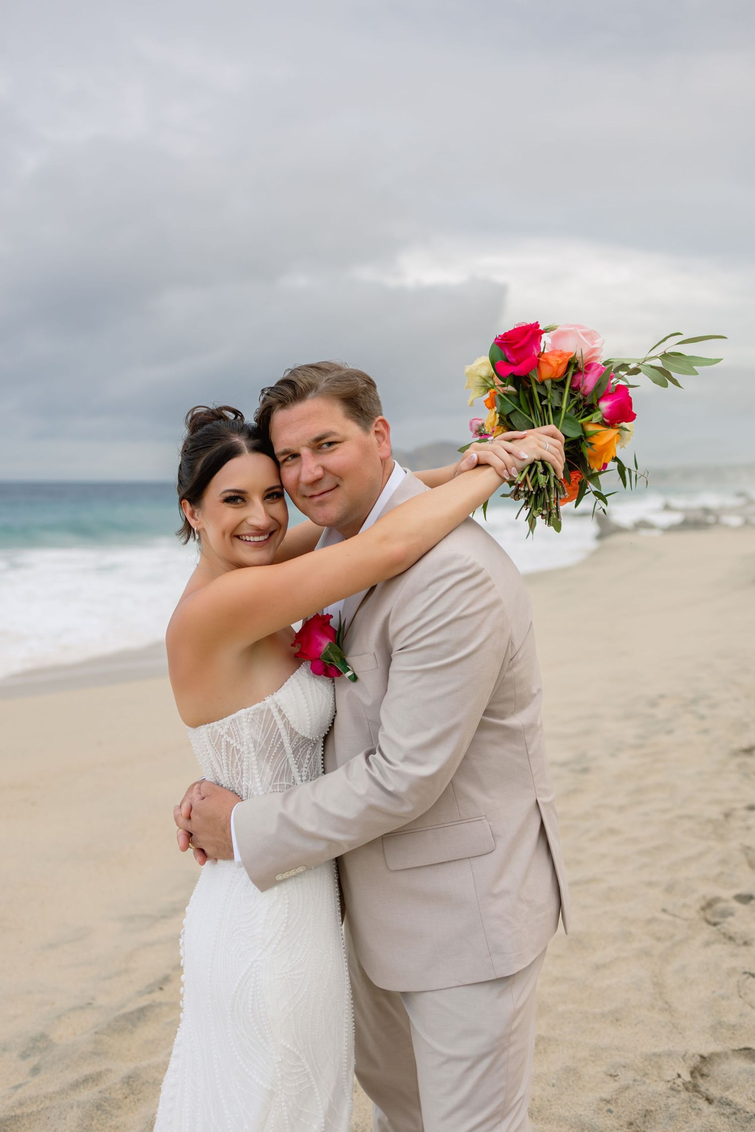 A couple embraces on a cloudy beach with colorful tropical flowers and white wedding attire.