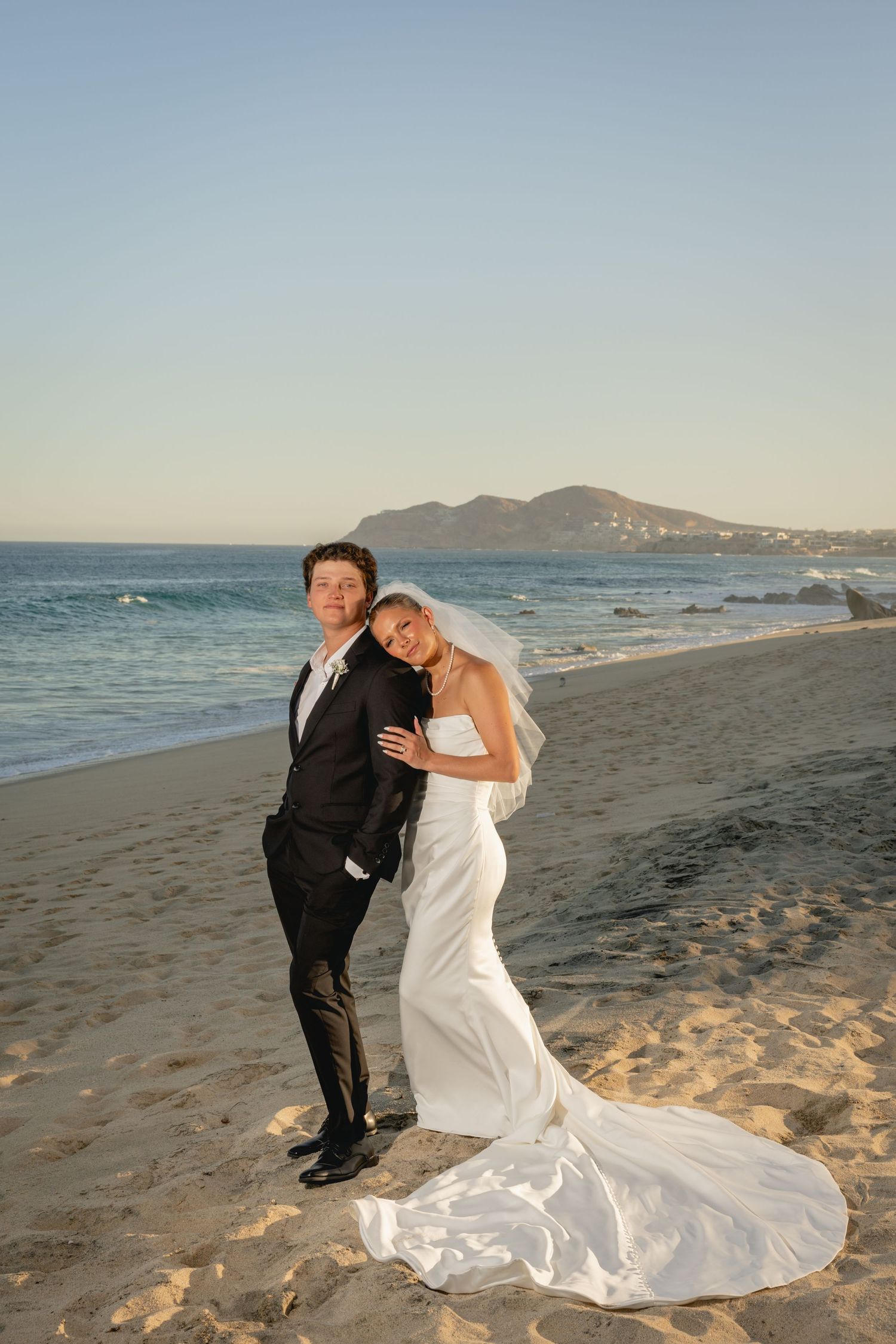 Bride and groom pose on a sandy beach at sunset, with ocean waves and a mountain in the background.