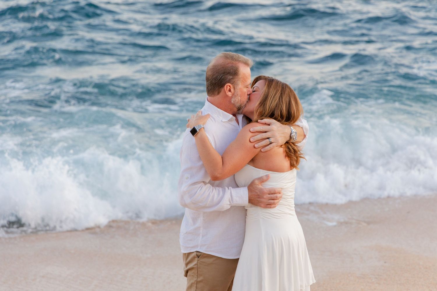 A couple shares a romantic kiss on a beach with crashing waves in the background during a sunset photo session.