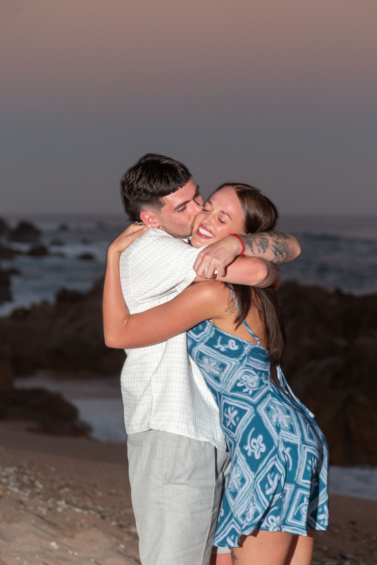 A romantic couple embraces on a beach at sunset with ocean and rocks in the background.