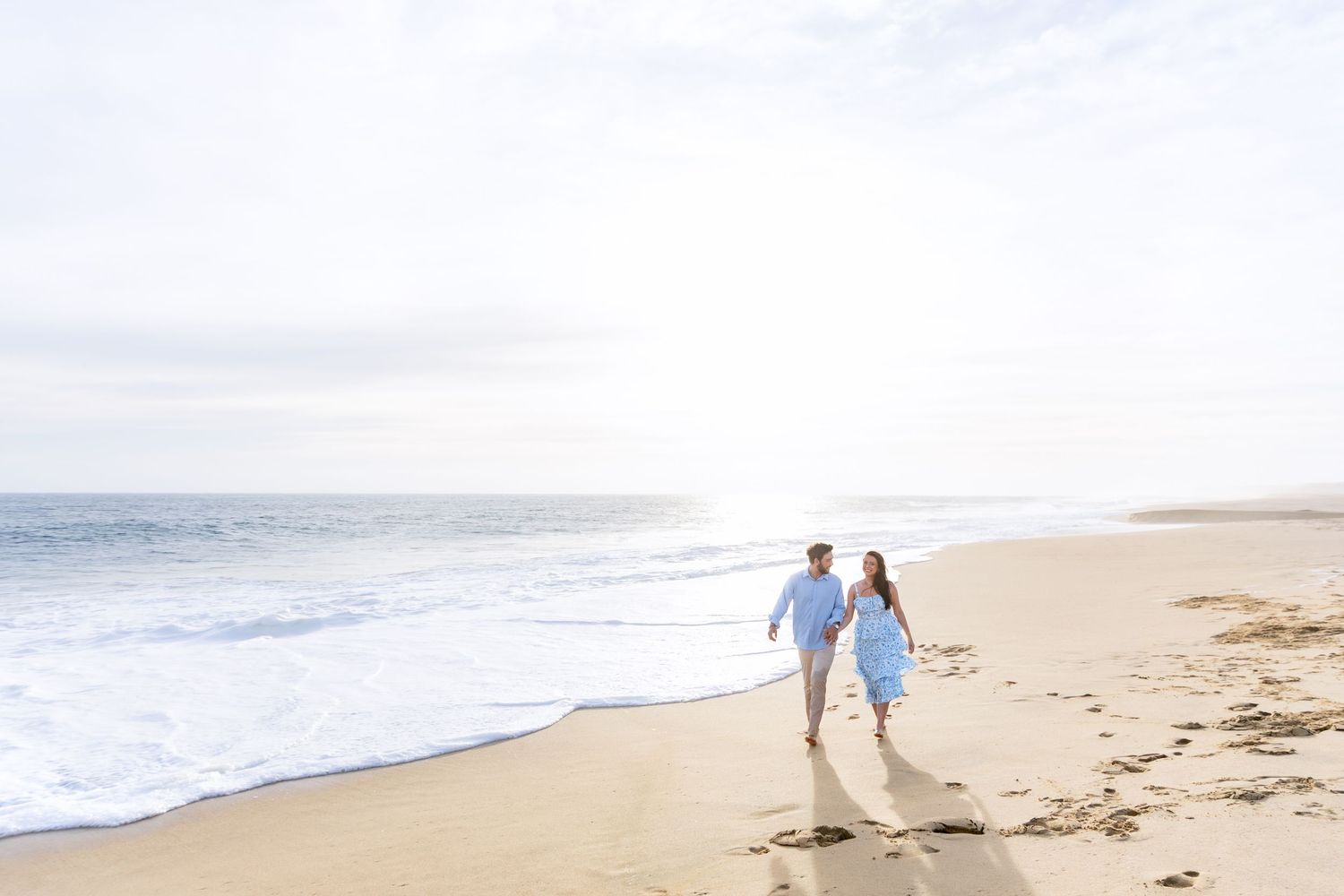 A couple in wedding attire walks along a misty beach shoreline with crashing waves in the background.