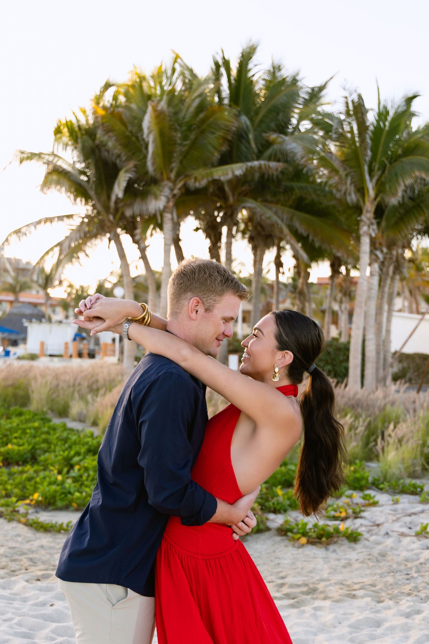 A romantic couple embraces on a tropical beach at sunset with palm trees swaying in the background.