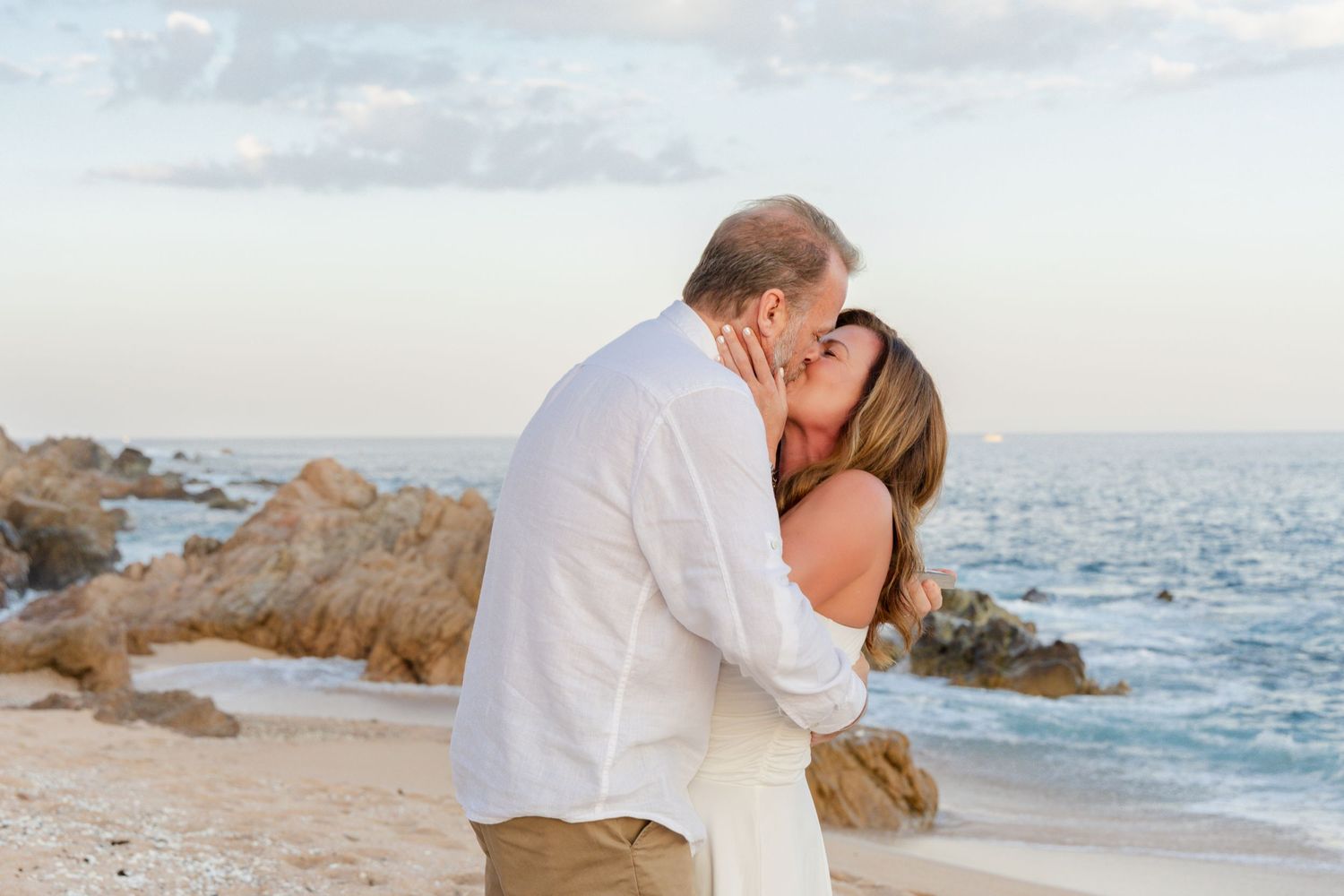 A romantic couple shares a kiss on a rocky beach at sunset with the ocean waves and cliffs in the background.