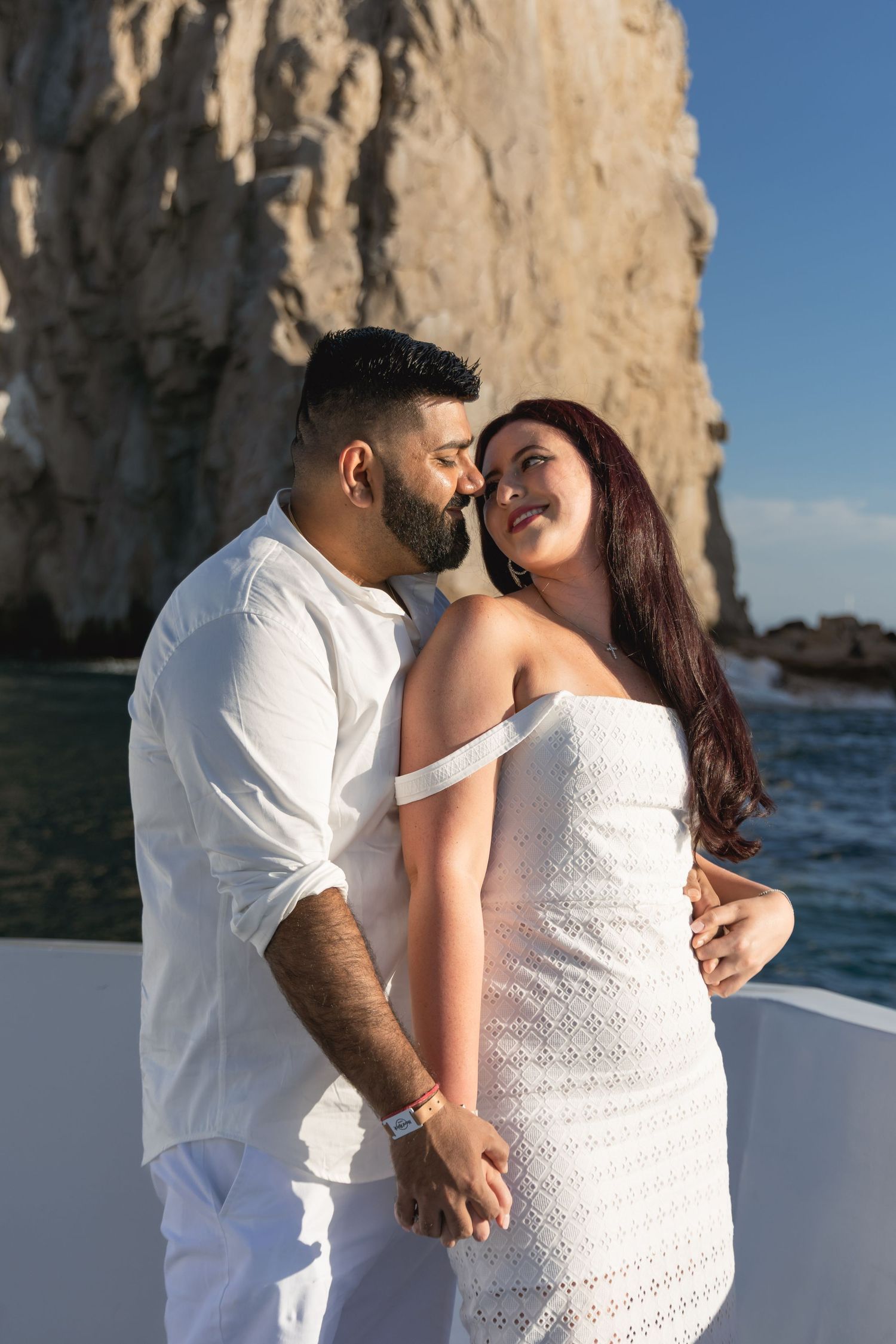 A romantic couple in white outfits share an intimate moment on a boat near dramatic coastal cliffs and blue water.
