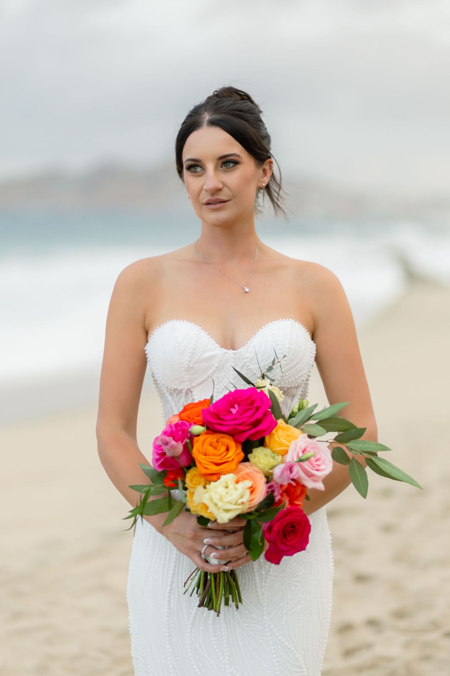 A bride in a white strapless gown holds a vibrant bouquet of pink, orange, and red flowers on the beach.