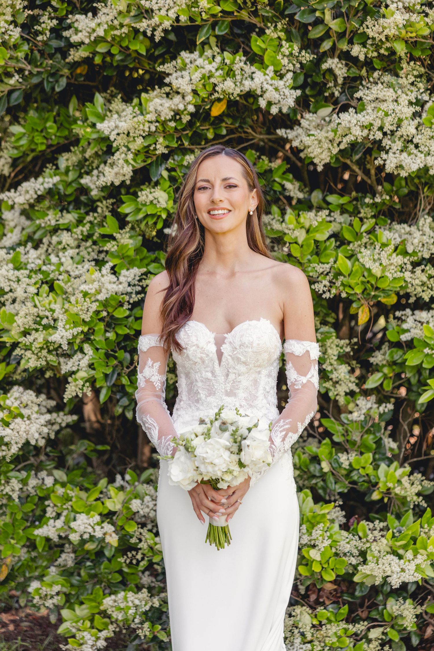Bride holding white bouquet against flowering bush backdrop.