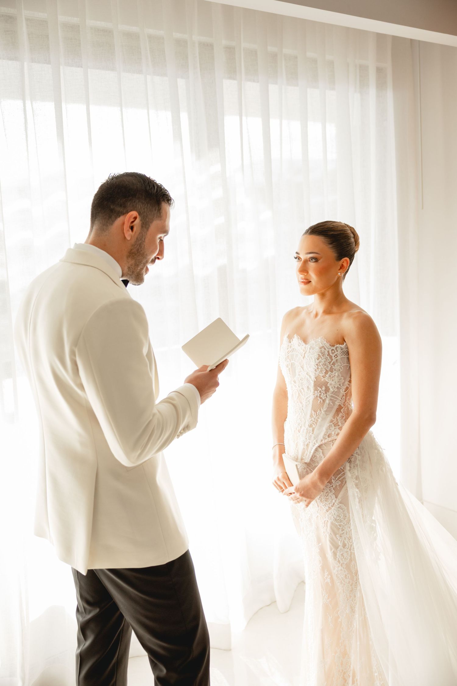 Wedding couple sharing a private moment while reading together near sheer curtains.